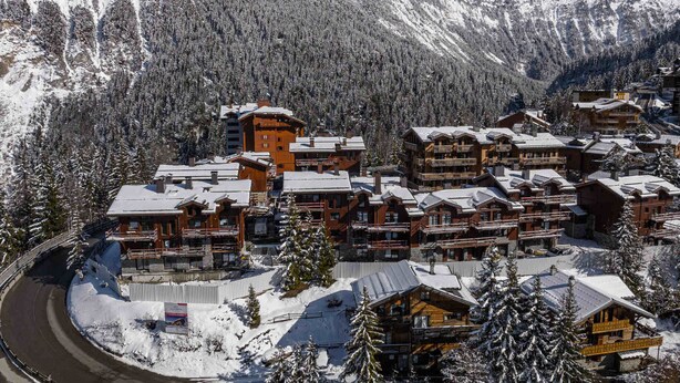 Um resort de montanha coberto de neve, com vários edifícios de madeira, incluindo chalés e um hotel, rodeado por florestas densas e montanhas.