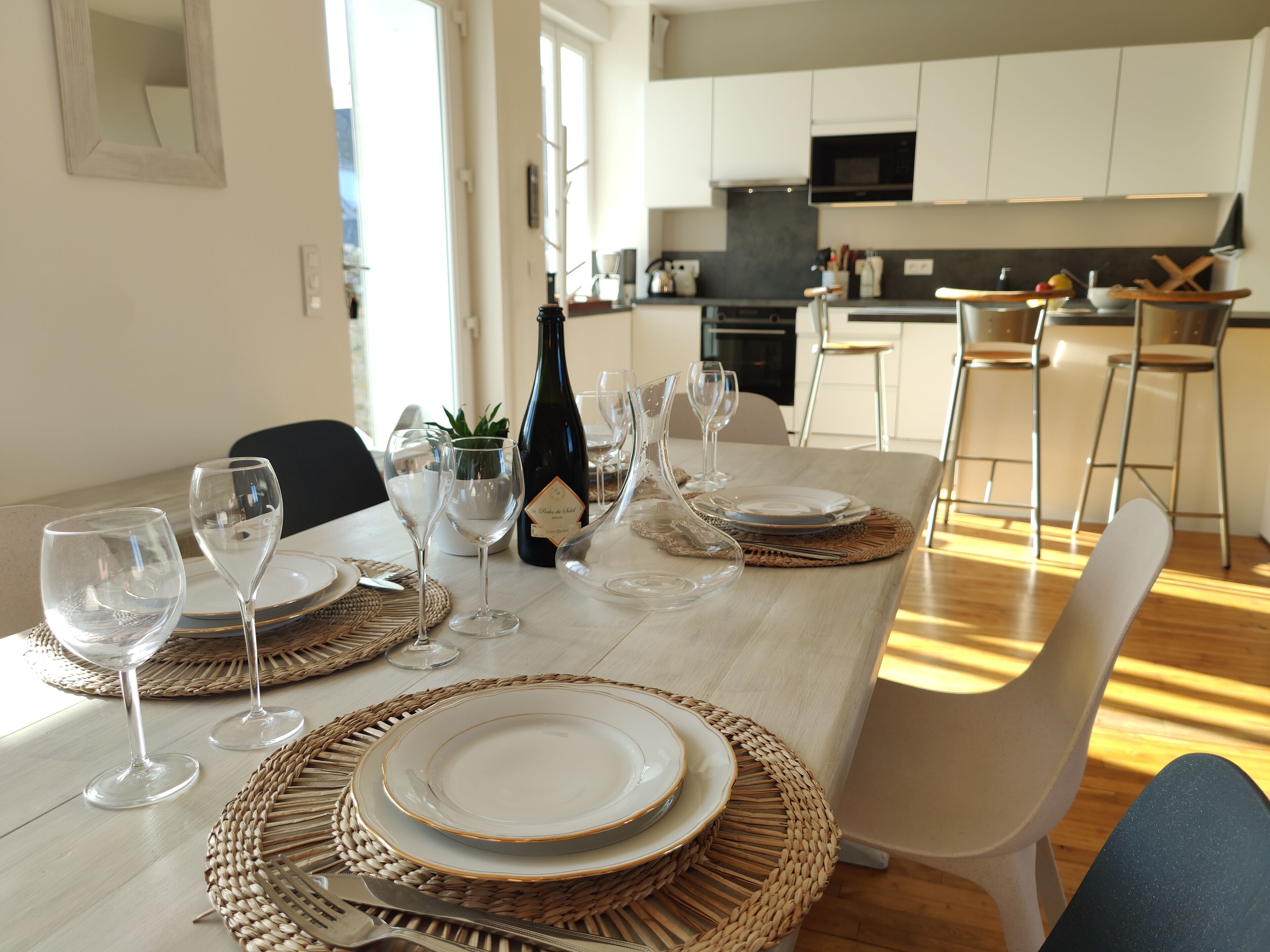 A dining table set with plates, glasses, and a bottle, with a kitchen in the background.