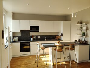 A modern kitchen with white cabinets, a black microwave, and a wooden countertop. There are three stools and a small table with a bowl of fruit. - Avel Dro House -  Pointe du raz - 4 peop. - 2 bathrooms - garden (Plogoff)