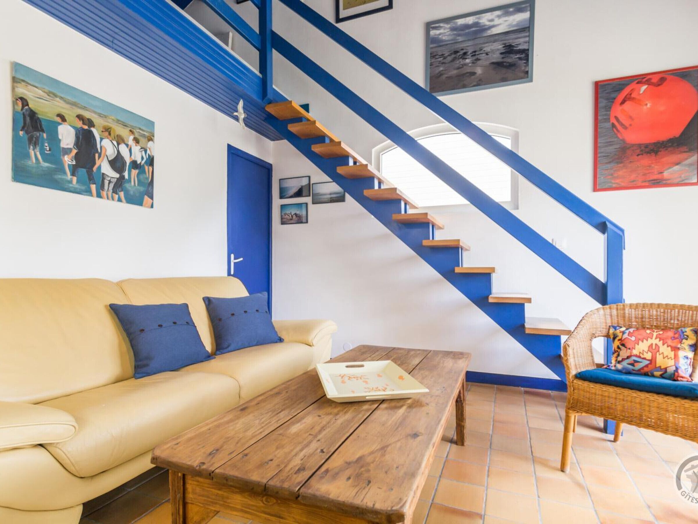 A living room with a beige sofa, a wooden coffee table, and a wicker chair. A blue staircase with wooden steps leads to a door.