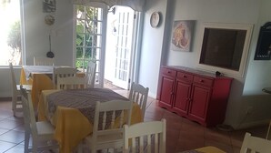 Un comedor con dos mesas, sillas blancas y manteles amarillos. Hay un aparador rojo y un televisor en la pared.