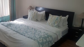 Una cama bien tendida con ropa de cama blanca y floral azul, un cabecero oscuro y una mesita de noche con una lámpara.