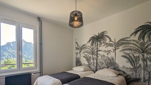 Un dormitorio con dos camas, una ventana con vistas a montañas, un radiador y una pared con un mural tropical.
