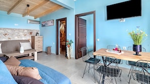 A living room with a sofa, a wooden dining table with chairs, a TV mounted on the wall, and an open door leading to another room. - Apartment 'Ventus Kalipso' with Sea View, Wi-Fi and Air Conditioning (Lefkada)
