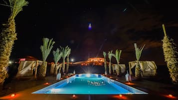 Un espace piscine aménagé pour la nuit, offrant une vue dégagée sur un bâtiment éloigné, des palmiers et un sol carrelé.