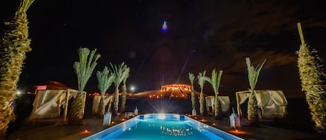 Un área de piscina nocturna con vista despejada a un edificio distante, palmeras y un piso de baldosas.
