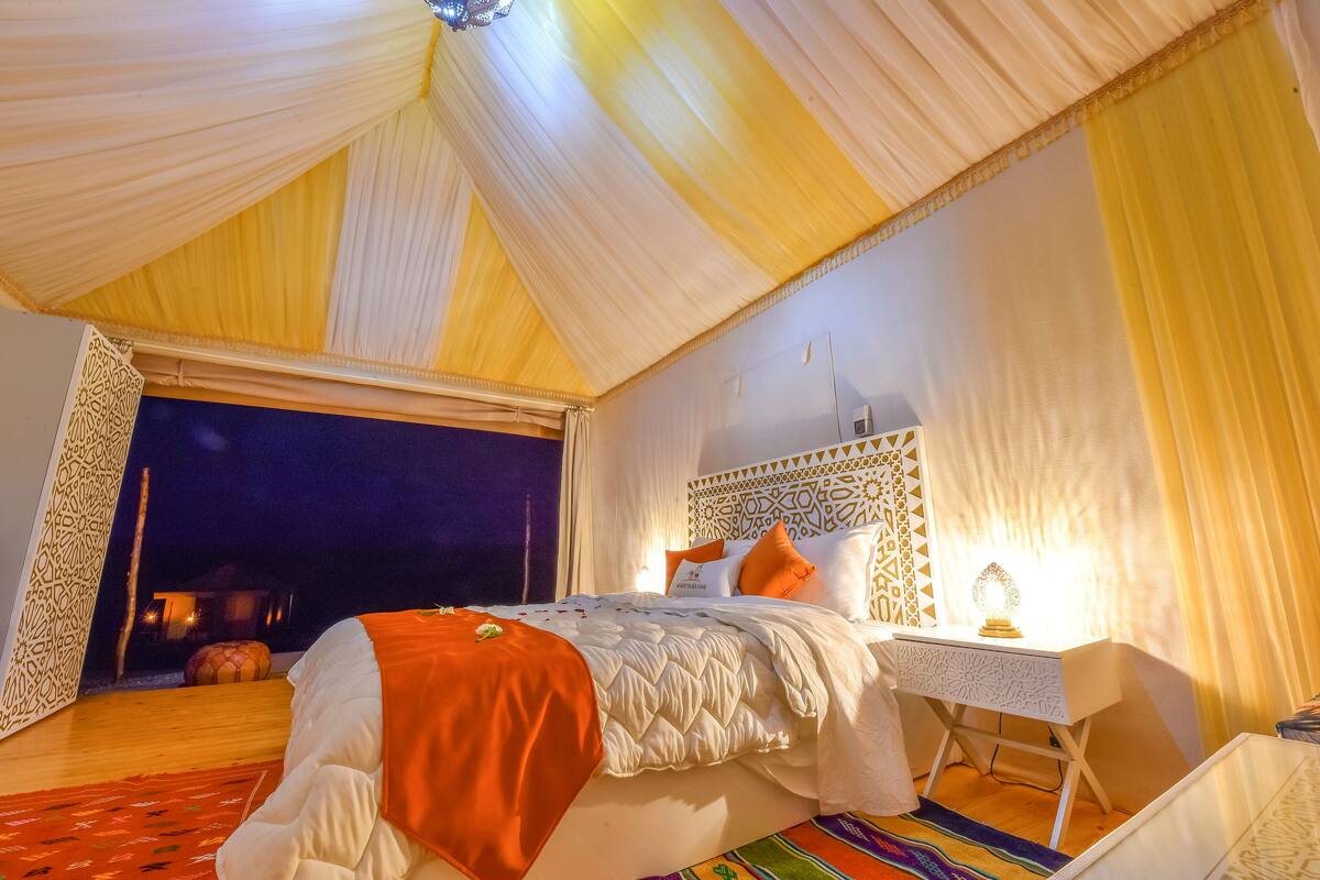 A room with a bed, a bedside table, and a nightstand. There is a window with curtains, a patterned wall, and a carpet.