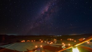 Cielo nocturno estrellado con una galaxia visible, un espacio al aire libre circular con valla de madera y luces colgantes.