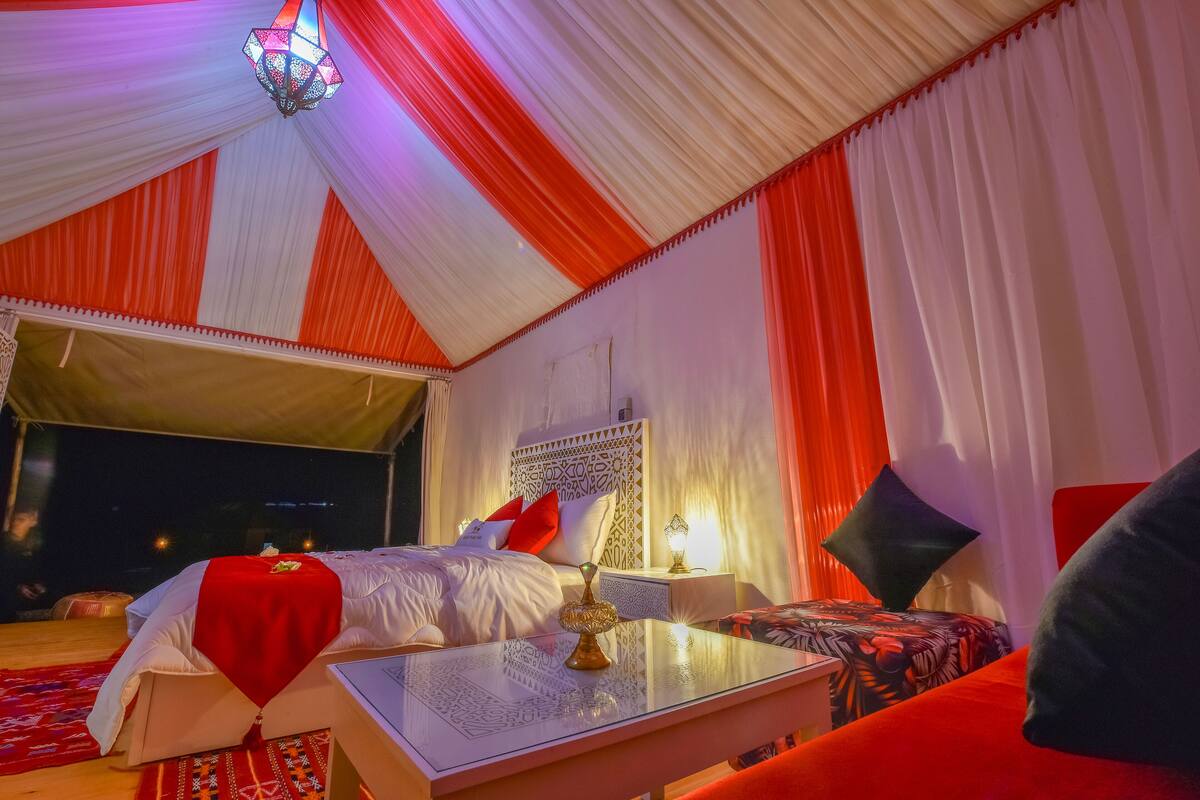A tented room with a bed, bedside table, and a view of the night sky.