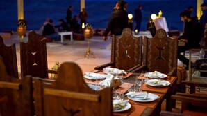 Comedor con mesas dispuestas para una comida, sillas y vista al mar de fondo.