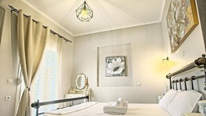 A hotel room with a bed, a bedside table, a nightstand, a wall-mounted light, a window with curtains, and a framed picture on the wall. - Apartment 'Ventus Artemis' with Sea View, Wi-Fi and Air Conditioning (Lefkada)