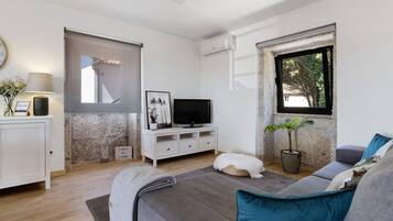 Un saló modern amb un sofà, una televisió sobre un moble blanc, una planta en test i una finestra amb vista a arbres.
