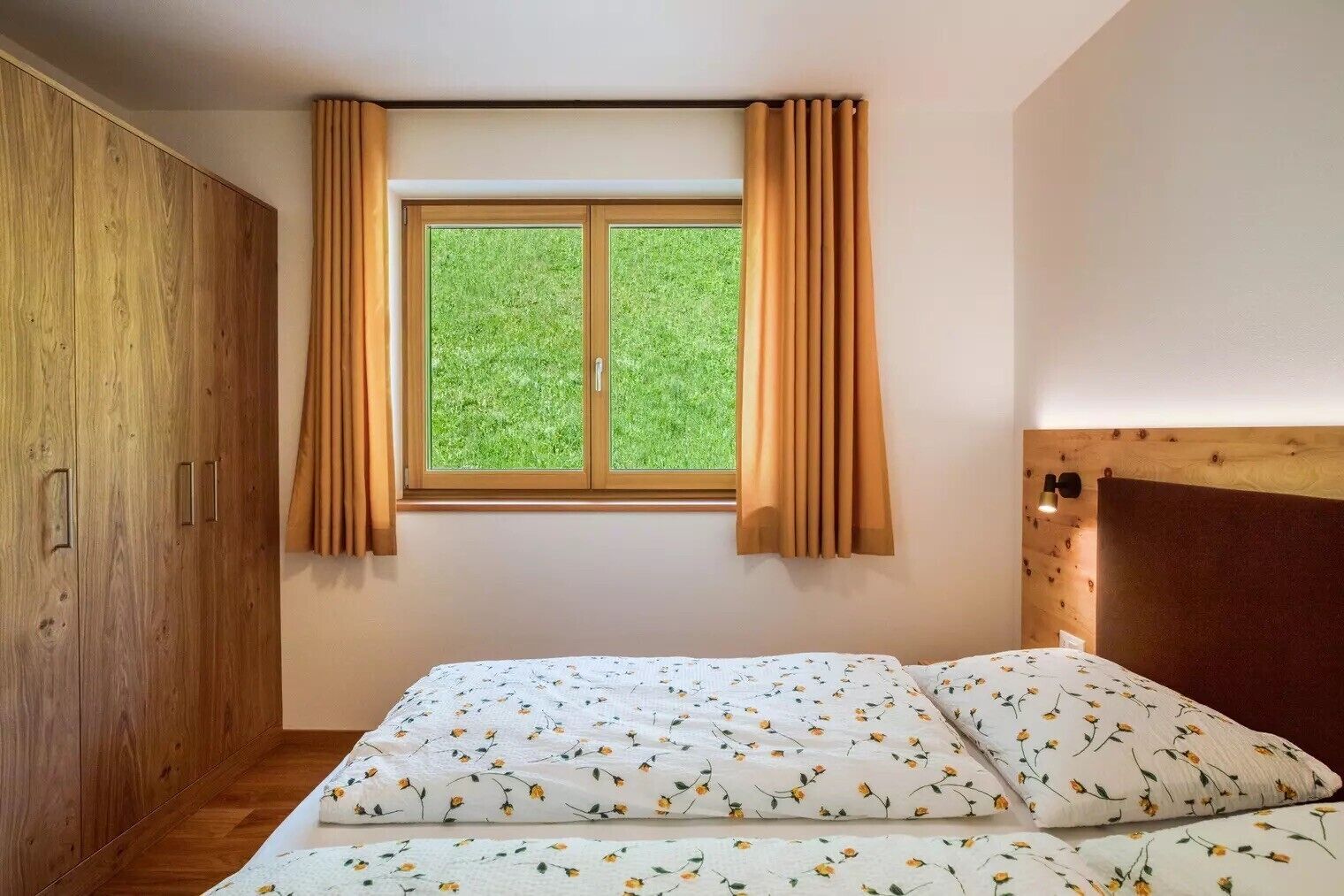 Un dormitorio con un armario de madera, una cama con ropa de cama floral, una ventana con cortinas y un cabecero con panel de madera.