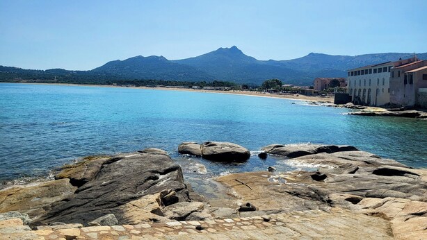 A coastal scene with a rocky foreground, a clear blue sea, and a mountain in the background.