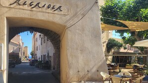 A narrow street with a stone archway labeled "Regione." There are outdoor tables and chairs under a canopy, suggesting a café or restaurant setting.