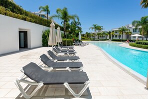Un área junto a la piscina con sillones de descanso y sombrillas, una piscina azul cristalina y un edificio blanco con una puerta.