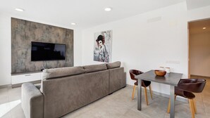 A modern living room with a sofa, a flat-screen TV mounted on a textured wall, a dining table with chairs, and a painting on the wall. - Vacation Home 'Suite Mirador Del Galeón' with Sea View, Wi-Fi and Air Conditioning (Adeje)