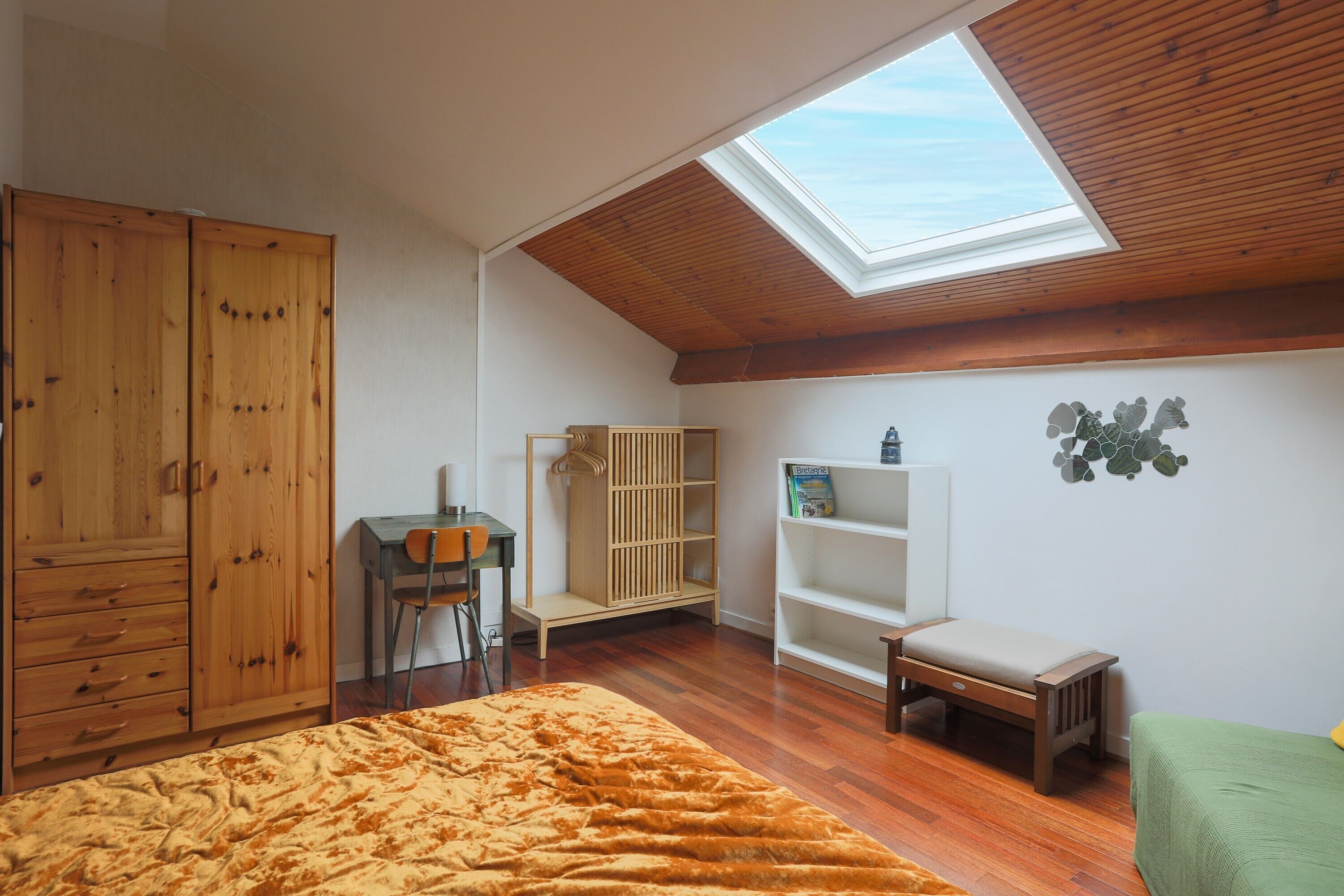 A bedroom with a skylight, wooden wardrobe, a desk, a bench, and a green bed.