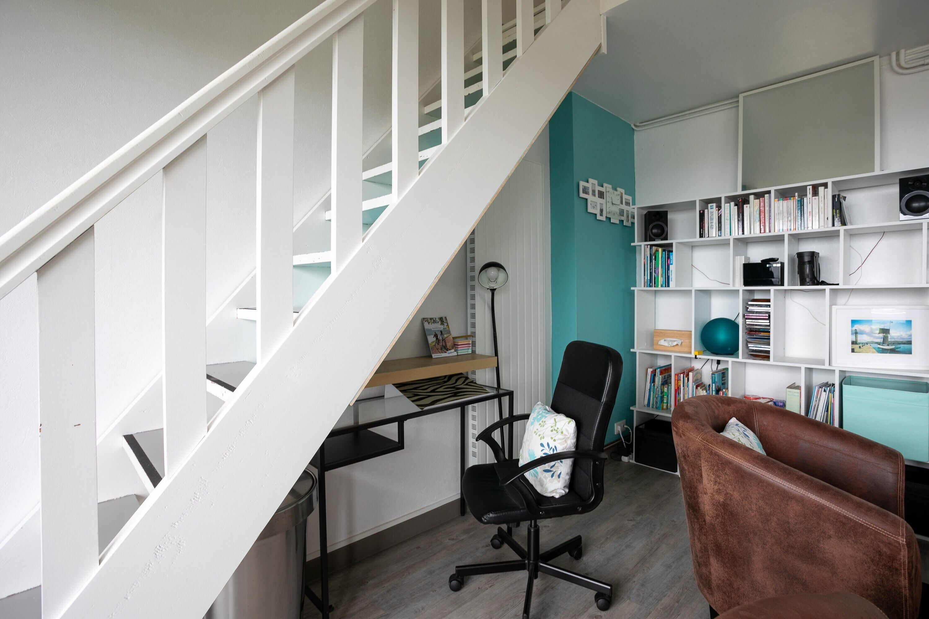 Un espace de bureau moderne avec un escalier blanc, un bureau avec un ordinateur, une chaise de bureau noire, un canapé marron et une bibliothèque remplie de livres et d’objets décoratifs.