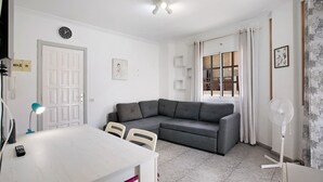 A room with a white door, a grey sofa, a dining table with chairs, a standing fan, and a window with curtains.