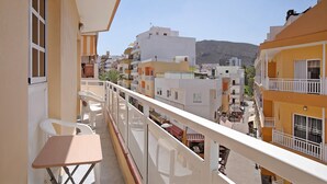 A balcony with a table and chairs, overlooking a street with buildings and a mountain in the background.