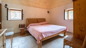 A bedroom with a wooden bed, a small wooden stool, a wicker chair, and two windows with metal bars. - Villa 'Venza with Pool' with Private Pool, Private Garden and Wi-Fi (Ostuni)