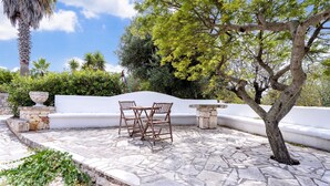 A stone patio with a white wall, two wooden chairs, and a tree. - Villa 'Venza with Pool' with Private Pool, Private Garden and Wi-Fi (Ostuni)