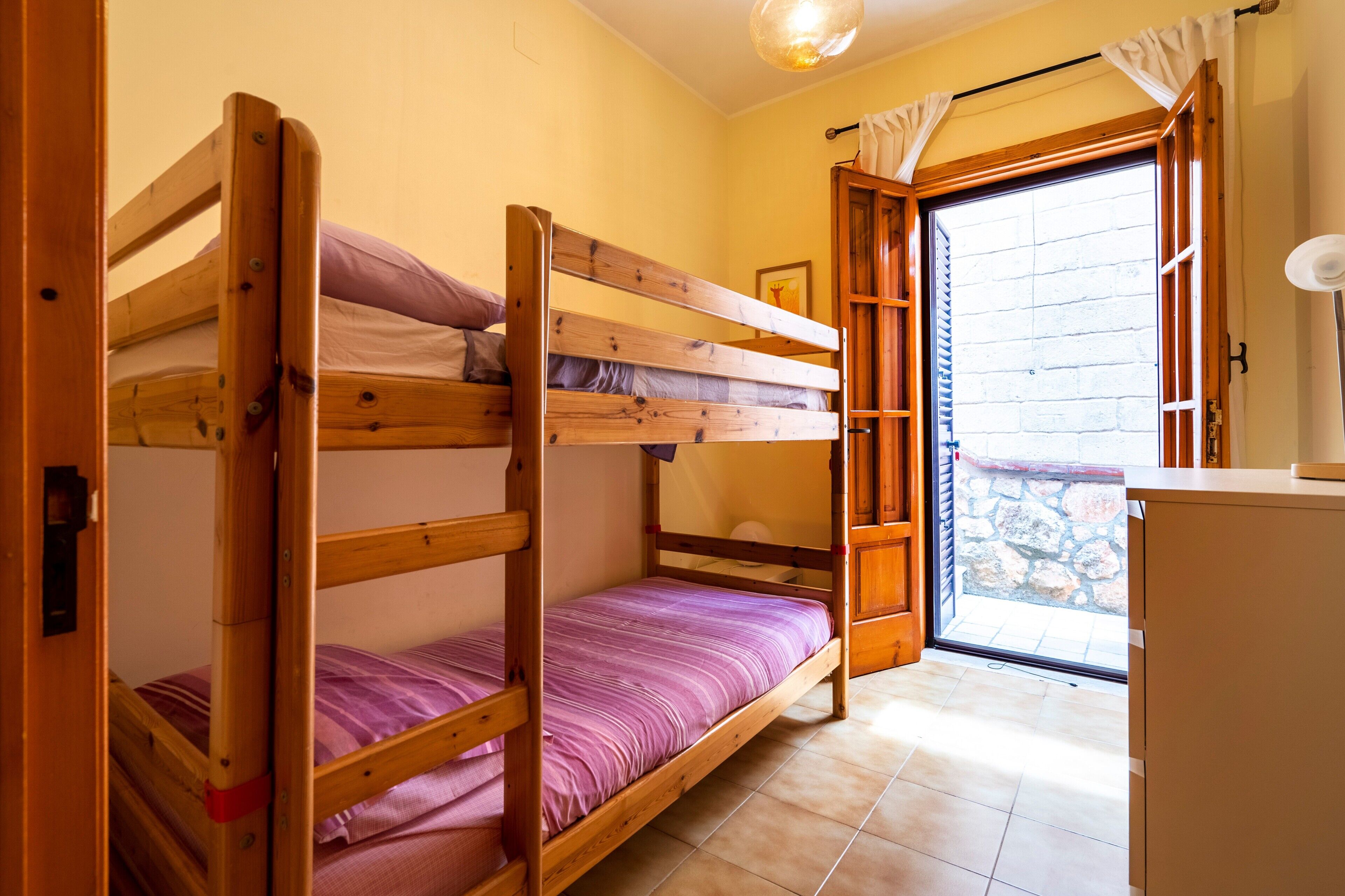 Ein Schlafraum mit Etagenbetten, einer Tür zu einem Balkon, einem kleinen Schreibtisch und einem Fenster mit Vorhängen.