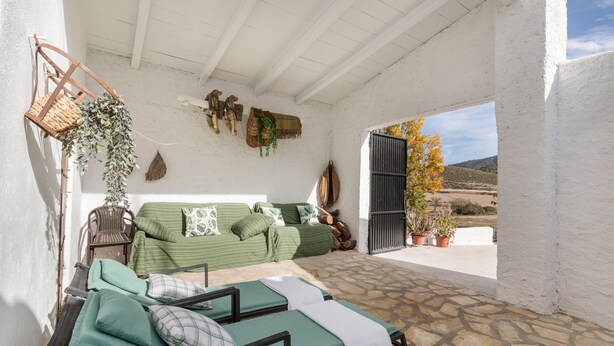 A covered outdoor seating area with a stone floor, a green sofa with cushions, and a wicker chair. - Holiday Home "Casa Rural Atalaya-Coy" with Mountain Views, Private Terrace and Garden (Coy)