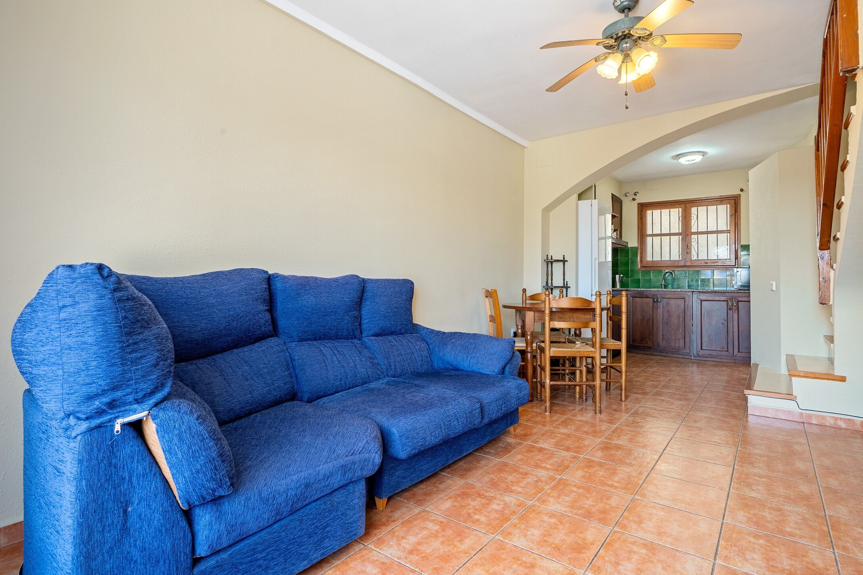 A blue sofa in a living area with a tiled floor, a ceiling fan, and a kitchen in the background.