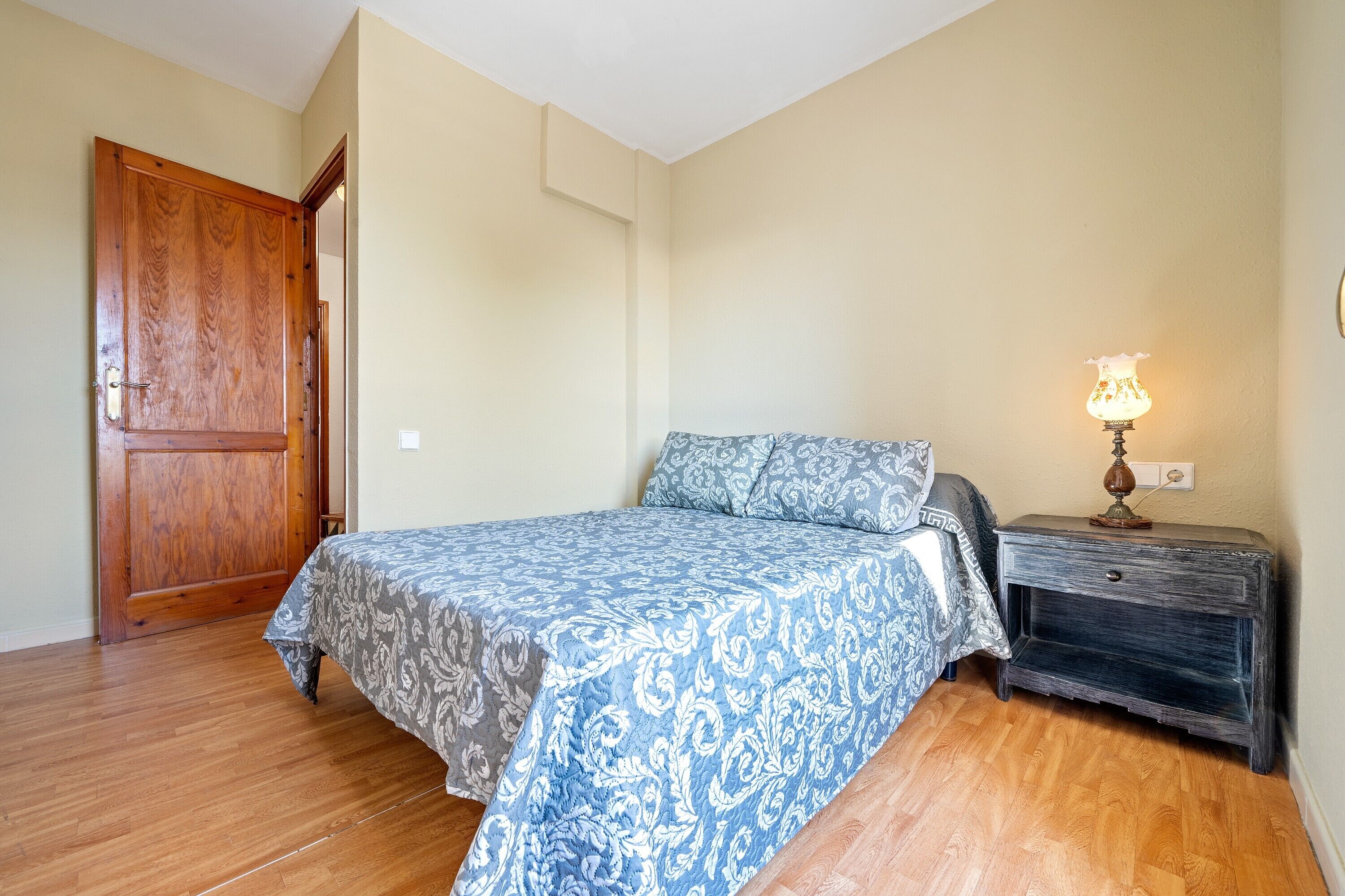 Un dormitorio con puerta de madera, cama con ropa de cama estampada, mesita de noche con lámpara y un enchufe de pared.