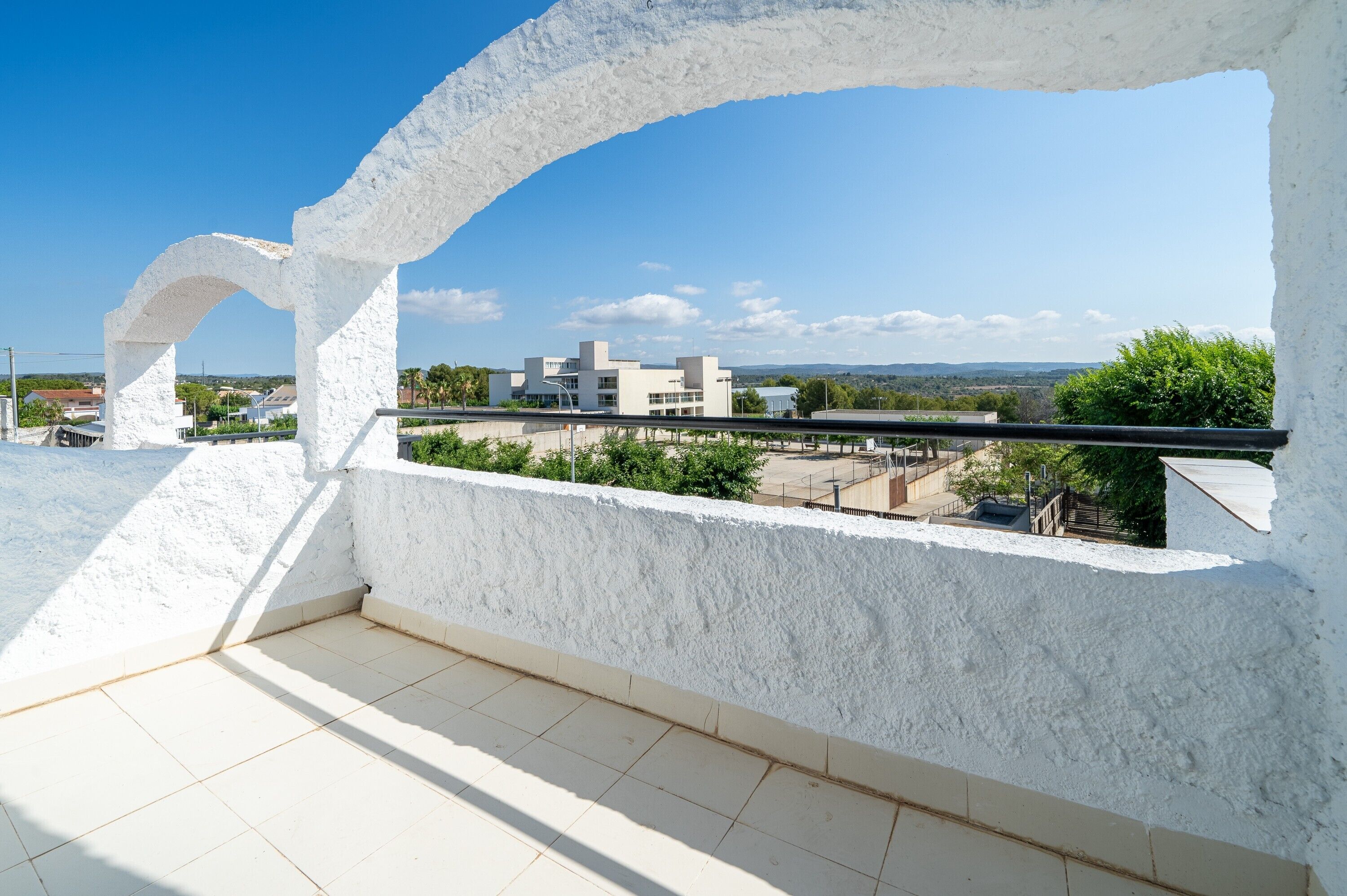 Un balcón con una pared blanca y con textura, piso de baldosas y vista a edificios y vegetación.