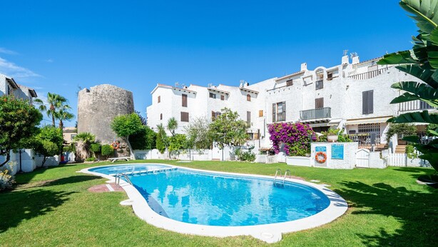 Una piscina rodeada por un césped cuidado, un edificio blanco con balcones y una alta torre de piedra.