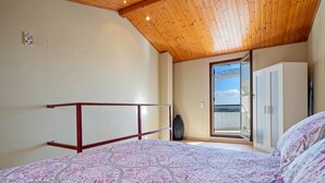 Un dormitorio con techo de madera, cama con ropa de cama estampada, un armario y vistas a través de un ventanal.