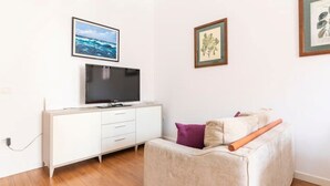 Ett modernt vardagsrum med en platt-TV på ett vitt skåp, en beige soffa med kuddar och två inramade tavlor på väggen.