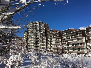 A multi-story residential building with balconies, surrounded by snow-covered trees and shrubs.