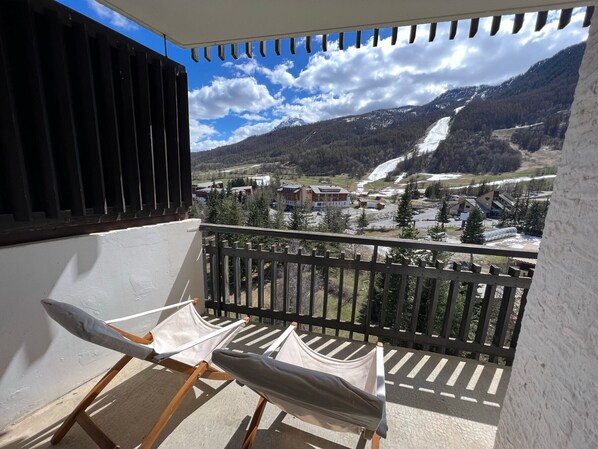 A balcony with two sun loungers, a view of a mountainous landscape, and a snow-capped peak in the distance.