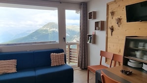 Un salon avec un canapé bleu, une table à manger avec des chaises, un téléviseur et une vue sur les montagnes par une grande fenêtre.