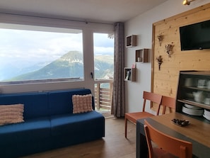A living room with a blue sofa, a dining table with chairs, a TV, and a view of mountains through a large window. - Comfortable studio cabin - Direct piste access, pets welcome (Plagne Aime 2000)