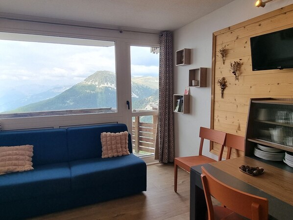 A living room with a blue sofa, a dining table with chairs, a TV, and a view of mountains through a large window.