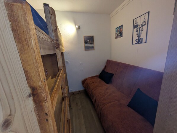 Un pasillo estrecho con una escalera de madera a la izquierda, un sofá marrón con un cojín negro a la derecha, y una pared con dos carteles: uno con la palabra 'Ski' y otro con una vista de montaña.