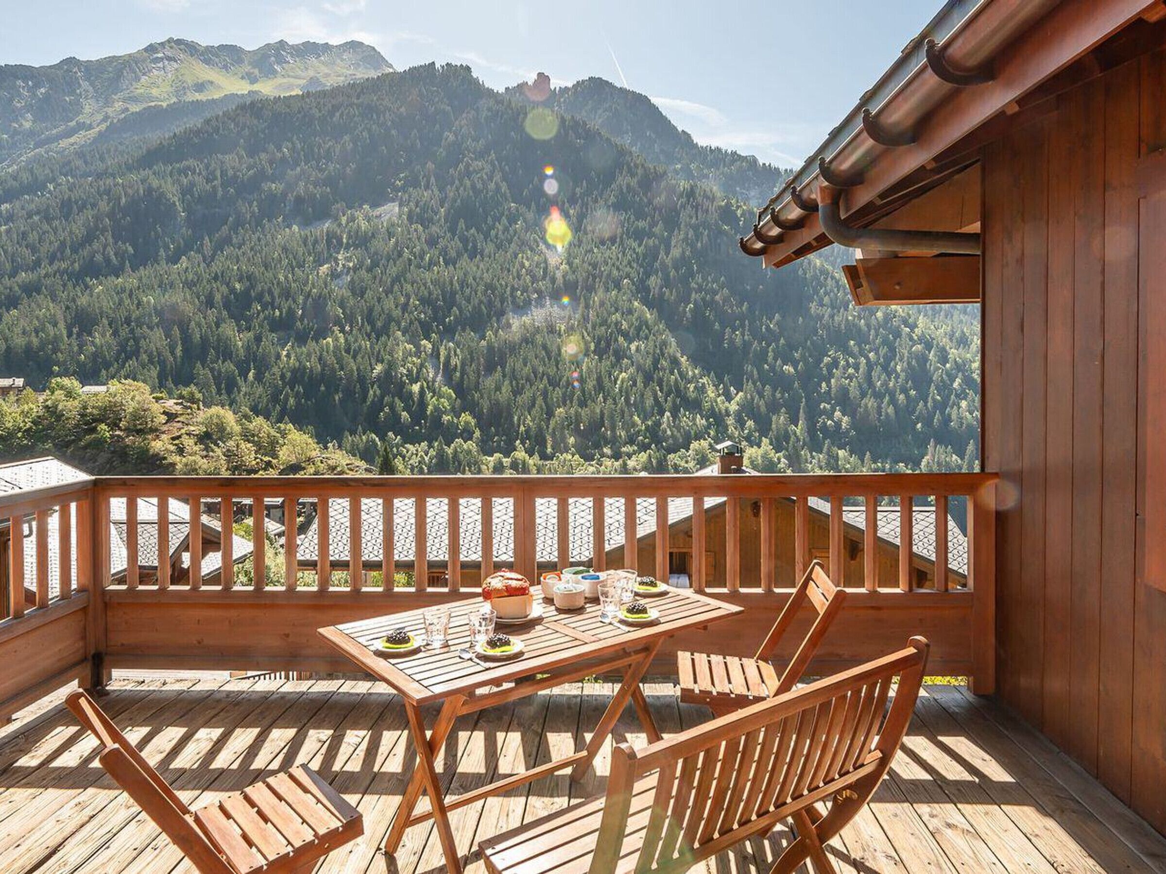 Eine Holzdielen-Terrasse mit Tisch und Stühlen, mit Blick auf einen bewaldeten Gebirgszug.