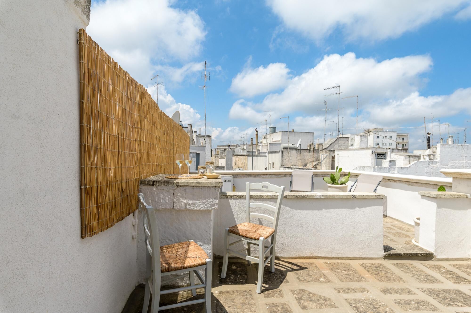 A rooftop terrace with a bamboo wall, two wicker chairs, and a small table with glasses.