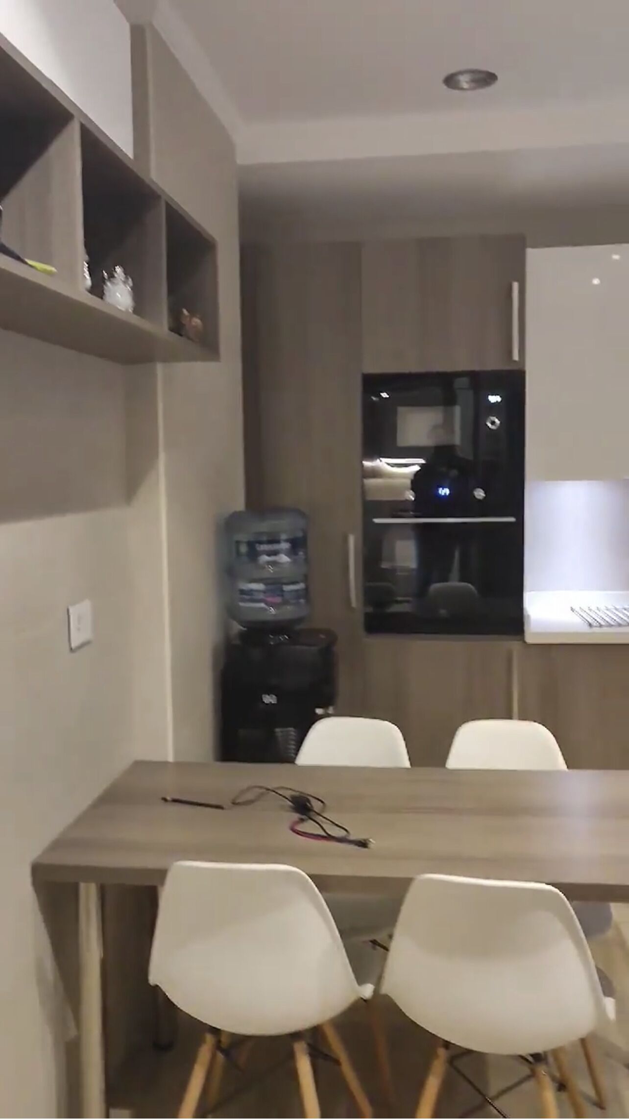 Une cuisine moderne équipée d’un réfrigérateur encastré, d’un distributeur d’eau et d’un four à micro-ondes. On aperçoit deux chaises blanches et une table au premier plan.