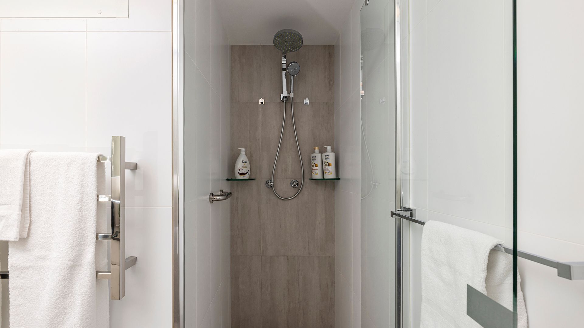 A modern shower with a handheld showerhead and a wall-mounted rain showerhead. There are two bottles of Dove body wash on a shelf.