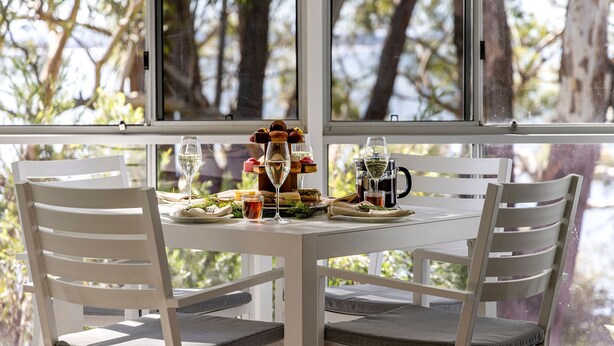 A dining table with white chairs, set with food and drinks, overlooking a body of water. - Belvedere Bayfront - the tranquillity of waterfront living (Nelson Bay)