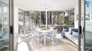 A modern dining area with a white table and chairs, a ceiling fan, and large windows offering a view of trees. - Belvedere Bayfront - the tranquillity of waterfront living (Nelson Bay)