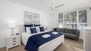 A bedroom with a bed, bedside tables, a chair, a ceiling fan, and a window with blinds. - Belvedere Bayfront - the tranquillity of waterfront living (Nelson Bay)