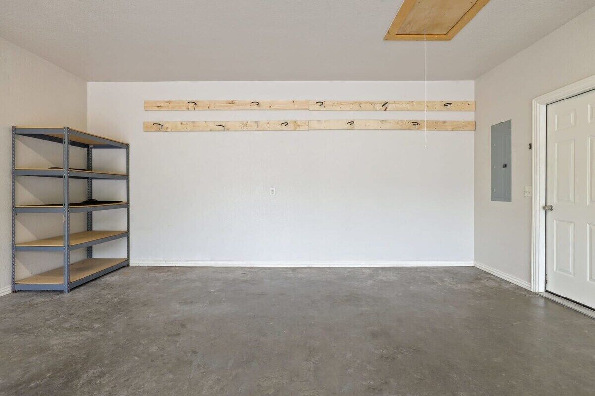 Una habitación con suelo de concreto, una estantería metálica, una puerta blanca y vigas de madera en el techo.