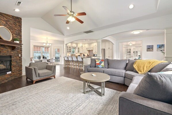 A spacious living room with a fireplace, a ceiling fan, and a kitchen in the background.